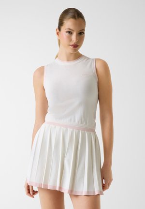 Młoda kobieta o blond włosach związanych w kucyk, ubrana w białą bezrękawnikową bluzkę oraz plisowaną białą spódnicę z różowym wykończeniem, stoi na jednolitym tle.