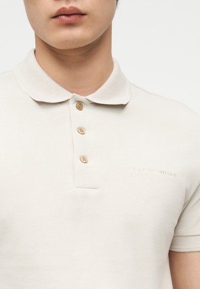 Εγγύτατη λήψη ενός ατόμου που φορά ένα μπεζ πόλο πουκάμισο με τρία καφέ κουμπιά και διακριτική κέντηση "Tiger of Sweden".