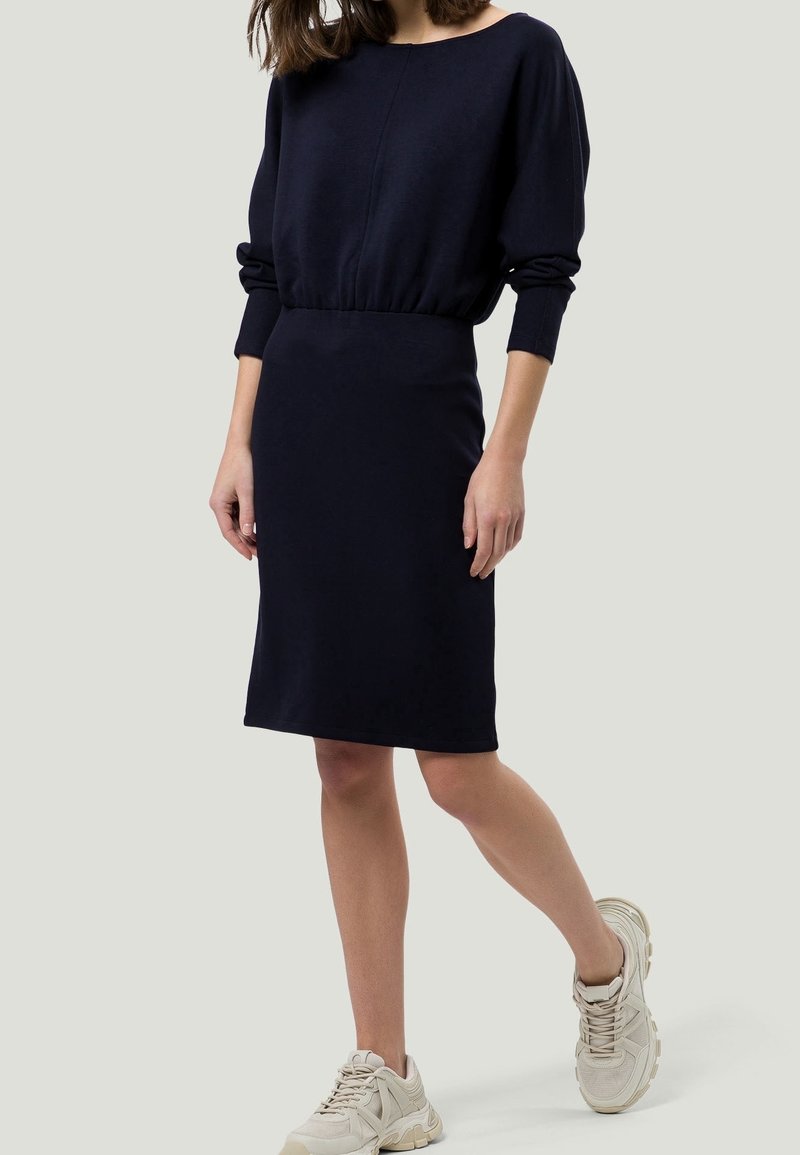 Femme portant une robe noire cintrée longueur genoux à manches longues et des baskets beiges, debout avec une jambe légèrement en avant.