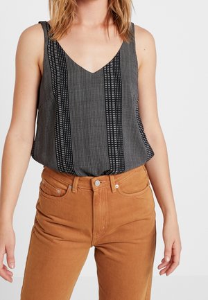 Donna che indossa un top senza maniche a righe bianche e nere infilato in un pantalone marrone a vita alta, in piedi davanti a uno sfondo semplice.