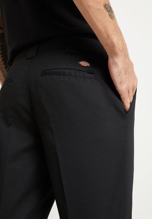 Close-up af en person iført sorte Dickies-bukser med hånden i lommen, der viser baglommen og en sommerfugletatovering på underarmen.