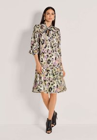Eine Frau trägt ein knielanges, langärmliges Satin Kleid mit lila, schwarz und beige Leopardenmuster, kombiniert mit schwarzen offenen Stiefeletten.