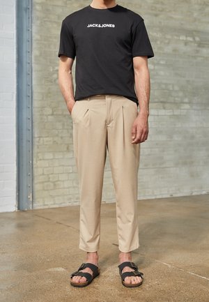 Man draagt een zwart Jack & Jones T-shirt, beige geplooide broek en zwarte sandaal met gesp, staand binnen op een betonnen vloer.