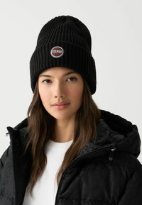 Črna rebrasta kapa z okroglim bordo rdečim logotipom z napisom "COLMAR", nošena s črno puhovko in belo srajco.