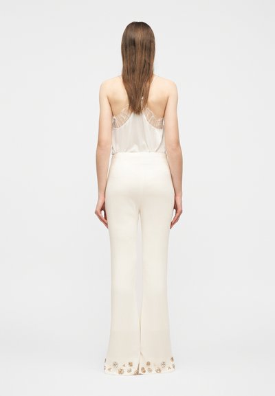 Γυναίκα με μακριά καστανά μαλλιά φοράει λευκό καλοκαιρινό τοπ με δαντέλα και κρεμ ψηλόμεσο παντελόνι με λουλουδένια διακοσμητικά στα φαρδιά μπατζάκια, γυρισμένη προς τα πίσω.