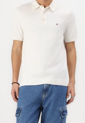 Mand iført en off-white kortærmet poloshirt med lille logo på brystet, kombineret med blå denim cargobukser og en ring på højre hånd.