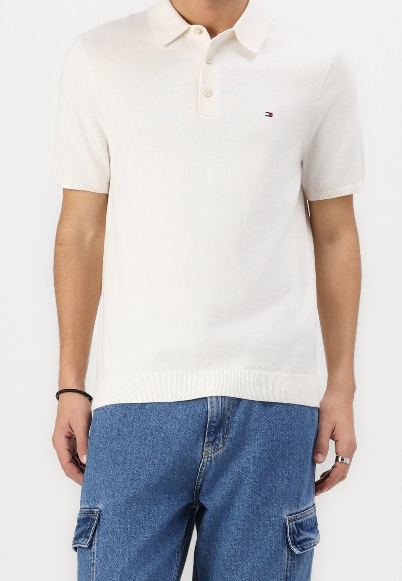 Homme portant un polo à manches courtes couleur blanc cassé avec un petit logo sur la poitrine, associé à un pantalon cargo en denim bleu et une bague à la main droite.