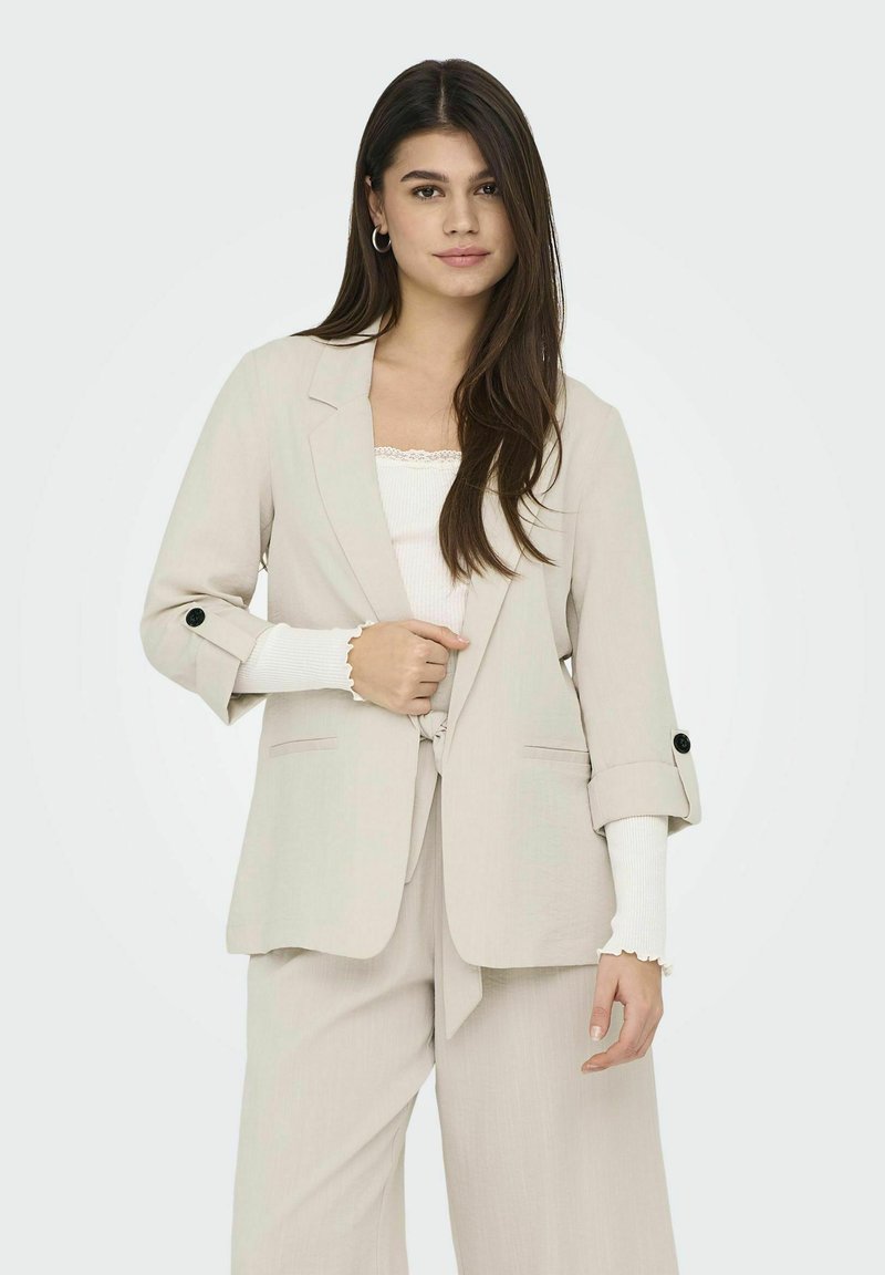 Hellbeige Blazer mit hochgerollten Ärmeln, schwarzen Knöpfen und einem Bindegürtel. Getragen über einem weißen Oberteil. Glatte Textur, taillierte Passform.