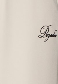 Λευκό κυλινδρικό αντικείμενο με τη λέξη "Pegador" γραμμένη σε μαύρη καλλιγραφική γραμματοσειρά στην επιφάνειά του.