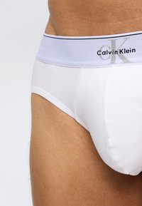 Αρσενικός κορμός που φοράει λευκά εσώρουχα Calvin Klein με ανοιχτό μπλε ζωνάκι στη μέση και μαύρα γράμμ ατα, πάνω σε απλό ανοιχτό φόντο.