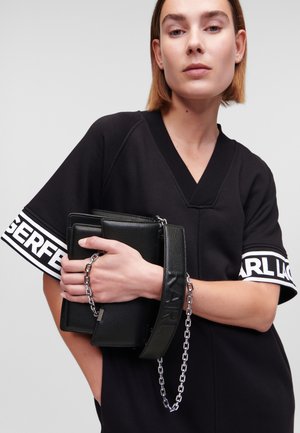 Clutch in pelle nera con superficie texture, dotato di una maniglia a catena. Il modello indossa una maglietta nera con logo bianco sulle maniche.