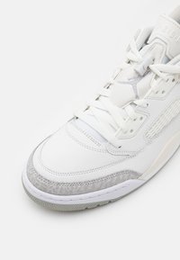 Valkoinen tennari, jossa on teksturoidut harmaat yksityiskohdat, sileää nahkaa ja rei'itetty muotoilu. Varustettu valkoisilla nauhoilla ja tyylitellyllä logolla kielen kohdalla.