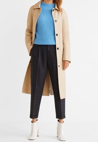 Beżowy trench coat z guzikami, niebieski prążkowany sweter, spodnie w kratę oraz białe botki za kostkę. Skup się na neutralnych i żywych kolorach, nowoczesnym kroju.