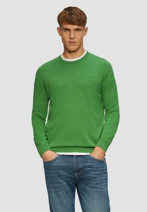 Grüner Baumwollpullover mit langen Ärmeln, gerippten Bündchen und Rundhalsausschnitt. Getragen über einem weißen T-Shirt, kombiniert mit blauen Jeans.