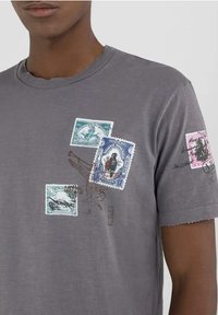 Camiseta de algodón gris de manga corta con cuatro impresiones de sellos vintage multicolores, incluyendo un dibujo de un pájaro y detalles de texto.