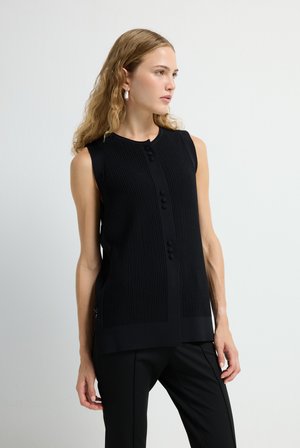 Zwarte mouwloze top met ribbelstructuur, ronde hals en een knoopsluiting aan de voorkant. Gepaard met zwarte geklede broek.