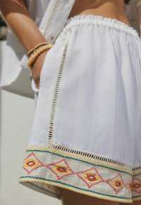 Vita bomullsshorts med broderade mönster i rött, gult och blått längs nederkanten och spetsdetaljer på sidorna. Elastisk midja.