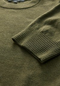 Oliven grøn striktrøje med en rund halsudskæring og ribstrikkede manchetter. Texturen er blød med en let fuzzy finish.
