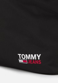 Czarna torba na ramię z wyraźnym białym i czerwonym logo "TOMMY JEANS". Tekstylna powierzchnia z solidną konstrukcją i delikatnym połyskiem.