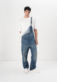 Denim overalls met een losse pasvorm, lichtblauw en donkerblauw kleurblokken ontwerp, verstelbare banden en zijknopen. Gecombineerd met een wit T-shirt en sneakers.