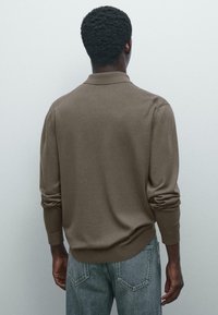 Mand med mørk hud iført en brun langærmet skjorte og lyseblå jeans står vendt mod en ensfarvet lys grå væg.