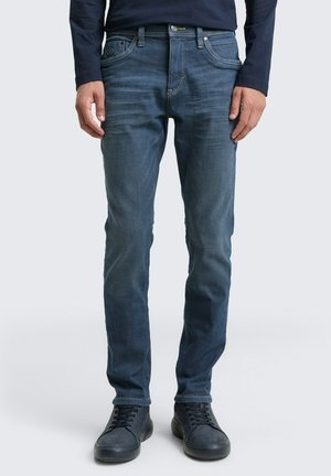 Homme portant un jean slim bleu et des baskets noires, debout devant un fond clair uni.