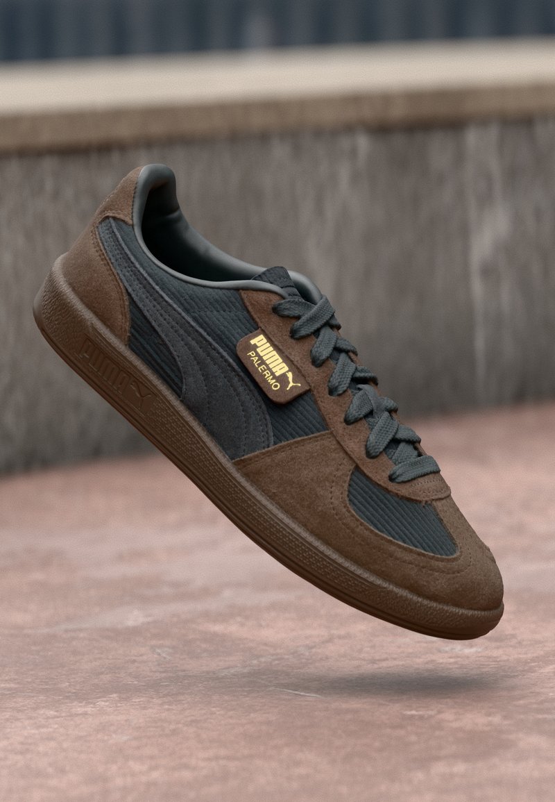 Basket Puma Palermo marron et noire avec des lacets gris, en l'air au-dessus d'une surface en béton et un arrière-plan flou.