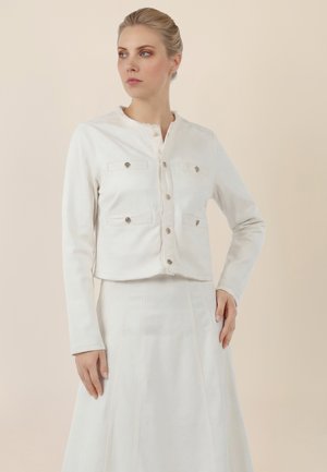 Woman wearing a white buttoned jacket with four front pockets and a matching white skirt, standing against a plain beige background.