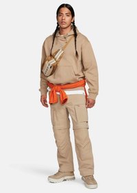 Beige hoodie med dragsnören, beige cargo byxor, orange bälte och en brun axelväska med vita ränder. Kraftiga beiga skor.