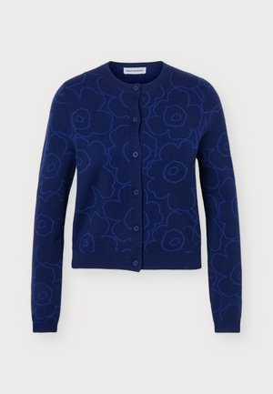 Femme portant un cardigan boutonné bleu foncé à motifs floraux, un jean bleu clair ample et des chaussures noires, debout devant un fond clair uni.