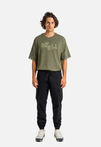 Junger Mann mit lockigem Haar, der ein olivgrünes, übergroßes T-Shirt mit dem Text "REELL", schwarze Cargo-Hosen und weiße Sneakers trägt, steht frontal.