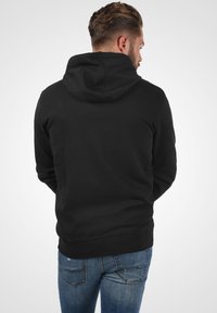 Zwarte pullover hoodie met een trekkoordcapuchon, zachte textuur en geribbelde zoom. Draagend met blauwe denim jeans, gezien vanuit de rug.