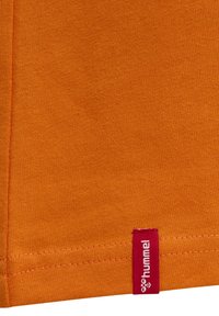 Orange bomullstyg med en slät textur har en röd etikett som är sydd vid fållen och visar varumärket "hummel."