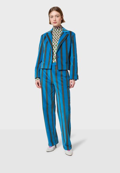 Dvodelni komplet koji se sastoji od plavog prugastog sakoa s uskim krojem i odgovarajućih hlača, uparen s uzorkovanim zeleno-bijelim rolkom.