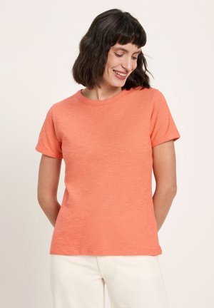 Frau mit kurzen dunklen Haaren, die lächelt und nach unten schaut, trägt ein einfaches oranges kurzärmeliges T-Shirt und weiße Hose, steht vor einem hellen Hintergrund.