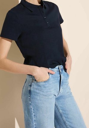 Persoon draagt een zwart poloshirt met korte mouwen in een lichtblauwe, hoog getailleerde spijkerbroek, met de handen in de voorzakken.