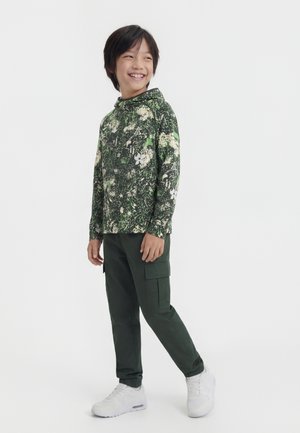 Copil zâmbitor în picioare, purtând un hanorac verde cu model, pantaloni cargo verde închis și adidași albi, pe un fundal alb.