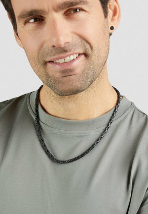 Mann trägt ein graues Rundhals-Shirt, schwarze Kettenkette und schwarzen Ohrstecker, lächelt mit leichtem Stoppelbart im Gesicht.