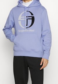 Felpa leggera viola realizzata in morbido cotone, con un grande logo grafico in nero e grigio. Include una tasca frontale e cappuccio con cordino.