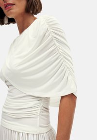 White dress features gathered fabric along the bodice and sleeves, creating a draped effect. Smooth texture with a fitted silhouette.