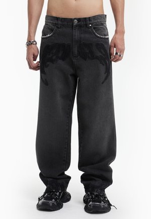 Personne portant un jean noir ample avec un motif de texte brodé foncé près des poches et des baskets noires, debout avec les bras détendus.