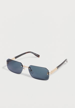 Suorakulmaiset tummansävyiset aurinkolasit, joissa on kultainen metallikehys ja mustat sankat, makaa valkoisella pinnalla.