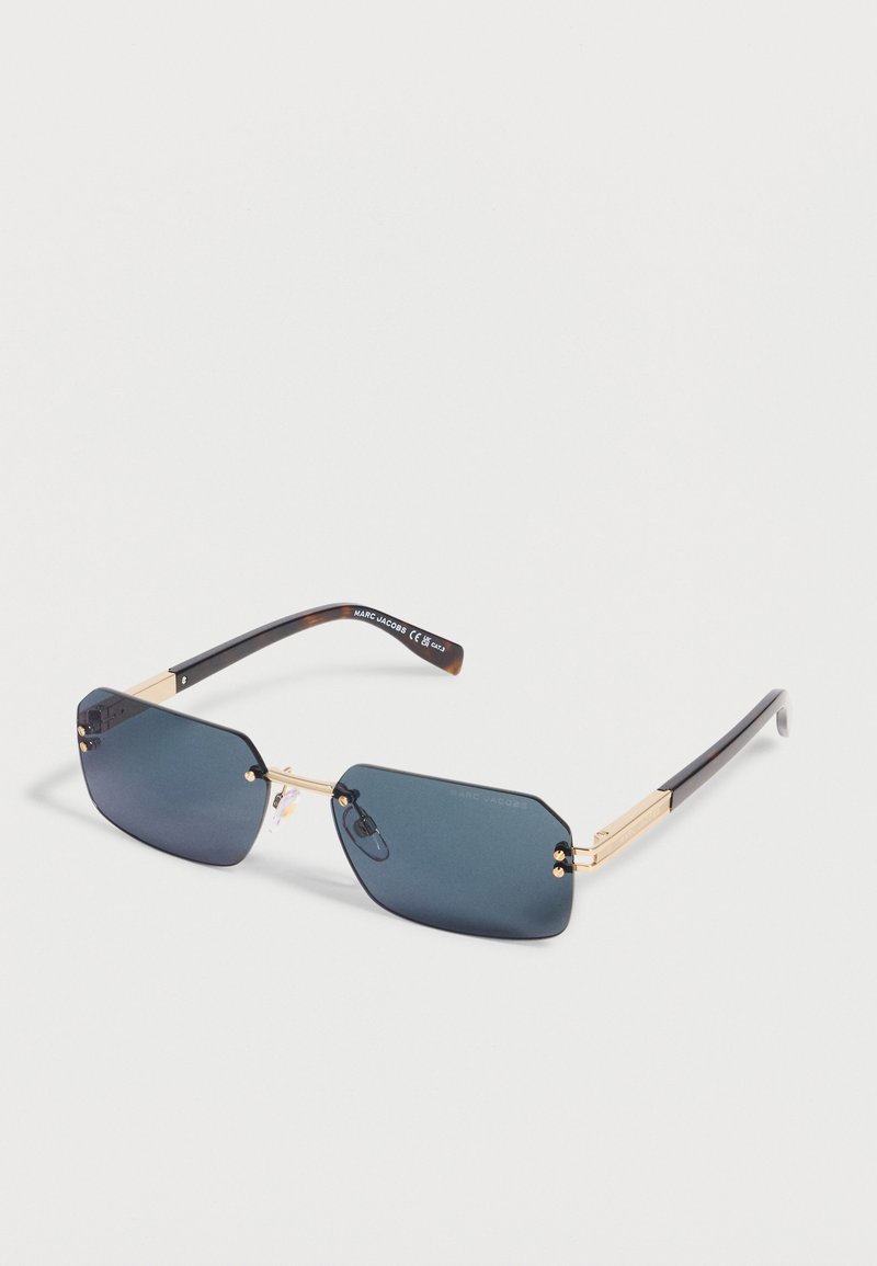 Ορθογώνια γυαλιά ηλίου με σκούρο φιμέ φακό, χρυσό μεταλλικό σκελετό και μαύρα μπράτσα, τοποθετημένα σε λευκή επιφάνεια.