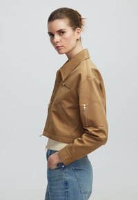 Braune cropped Jacke mit Kragen, Reißverschlusstaschen und Details aus goldenem Metall. Getragen mit blauen Jeans, die eine glatte Textur und eine taillierte Passform aufweisen.