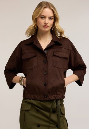 Blonde vrouw draagt een donkerbruine jas met knopen en klepzakken en een olijfgroene rok met knopen en zijzakken, handen in de jaszakken.