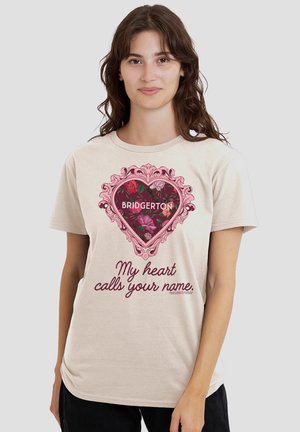 Young woman wearing beige T-shirt with pink ornate heart frame, floral pattern, "BRIDGERTON" text, and "My heart calls your name" script.