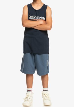 Person stående med armene over kors iført en sort Quiksilver tanktop, blå shorts, hvide sokker og hvide sneakers på en hvid baggrund.