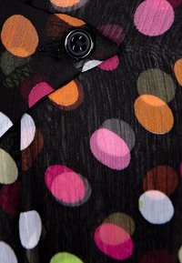 Schwarzer Stoff mit bunten Punkten in Pink, Orange, Grün und Weiß; verfügt über ein schwarzes Knopfdetail. Strukturierte, durchsichtige Optik.