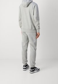 Man i ljusgrå hoodie och matchande joggare med svarta och vita Nike-sneakers, stående framför en neutral ljus bakgrund.