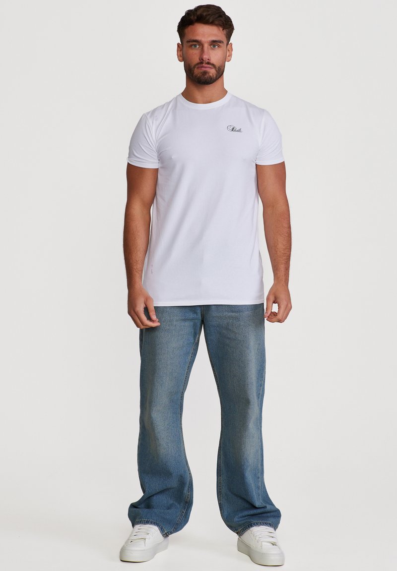Άνδρας με γενειάδα φοράει εφαρμοστό λευκό t-shirt με μικρό μαύρο λογότυπο στο στήθος, φαρδιά μπλε τζιν και λευκά αθλητικά παπούτσια, στέκεται σε μονόχρωμο φόντο.
